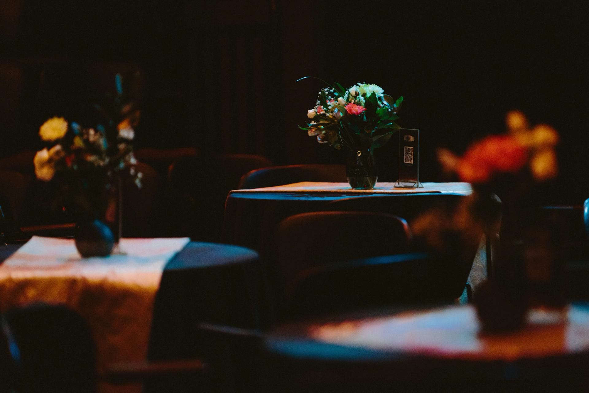 Dimly lit round tables with floral centrepieces and a small QR code sign.