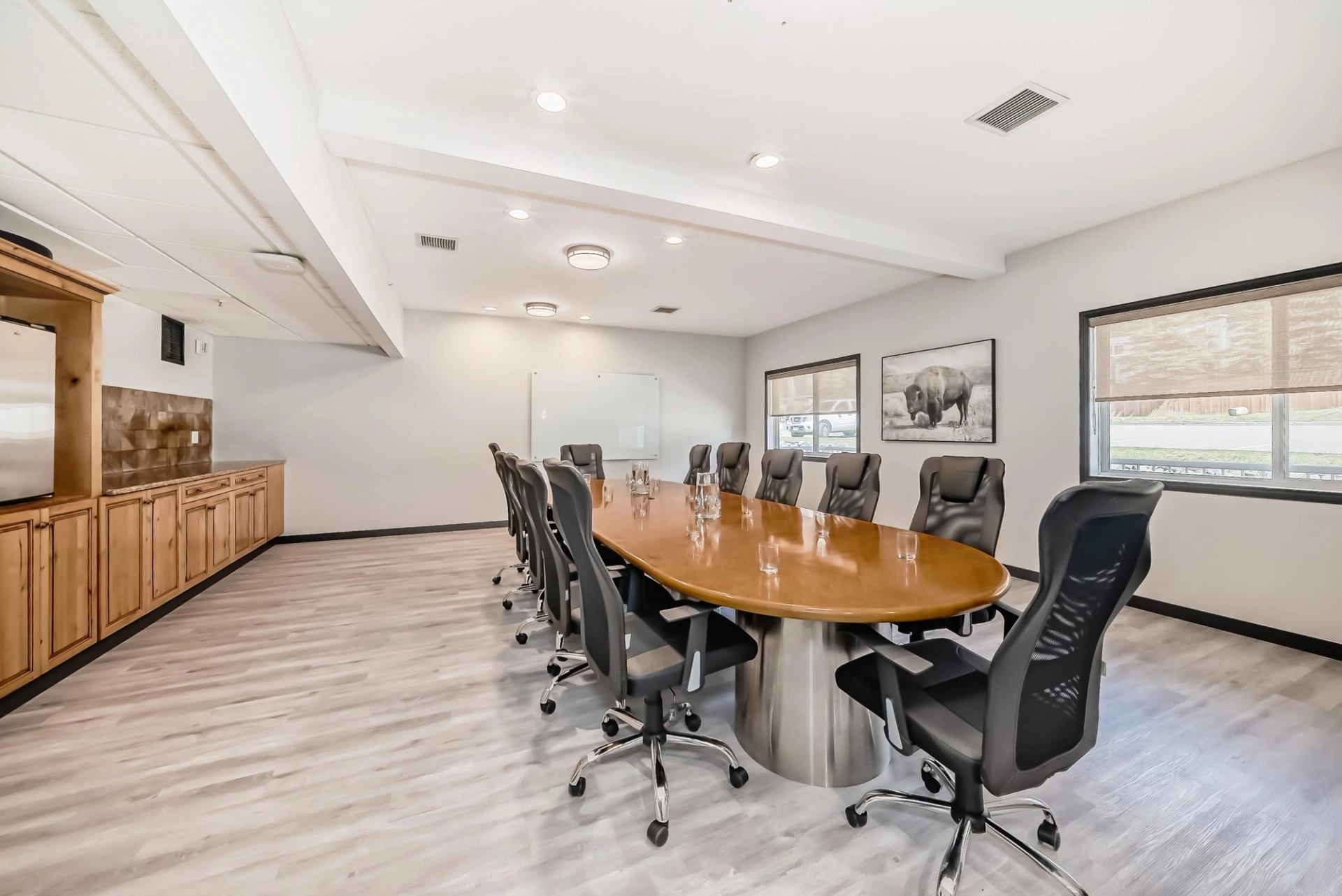 A well-lit conference room with a large oval table, several black office chairs, two windows, a whiteboard, wall art of a buffalo, and wooden cabinets.