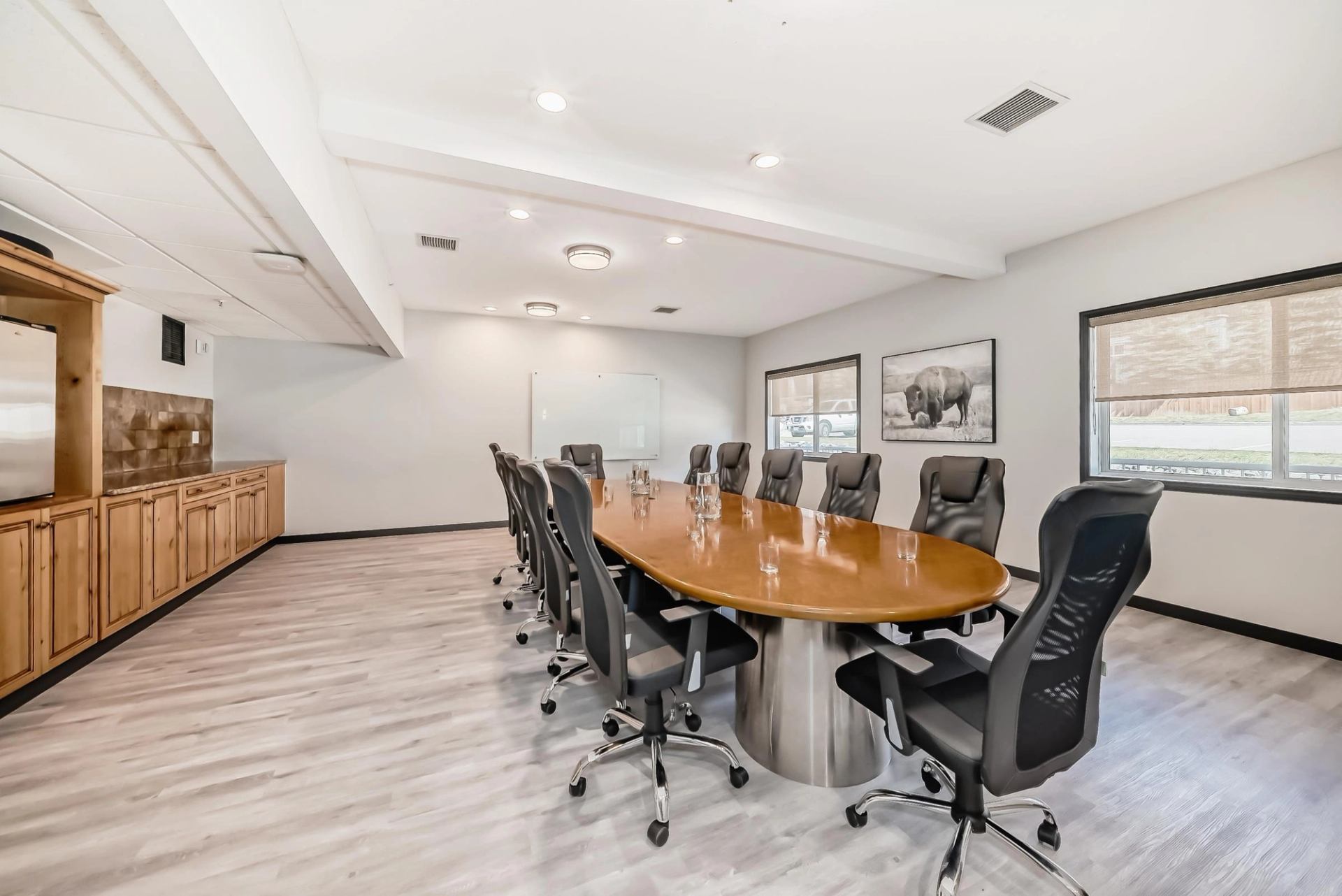 A well-lit conference room with a large oval table, several black office chairs, two windows, a whiteboard, wall art of a buffalo, and wooden cabinets.