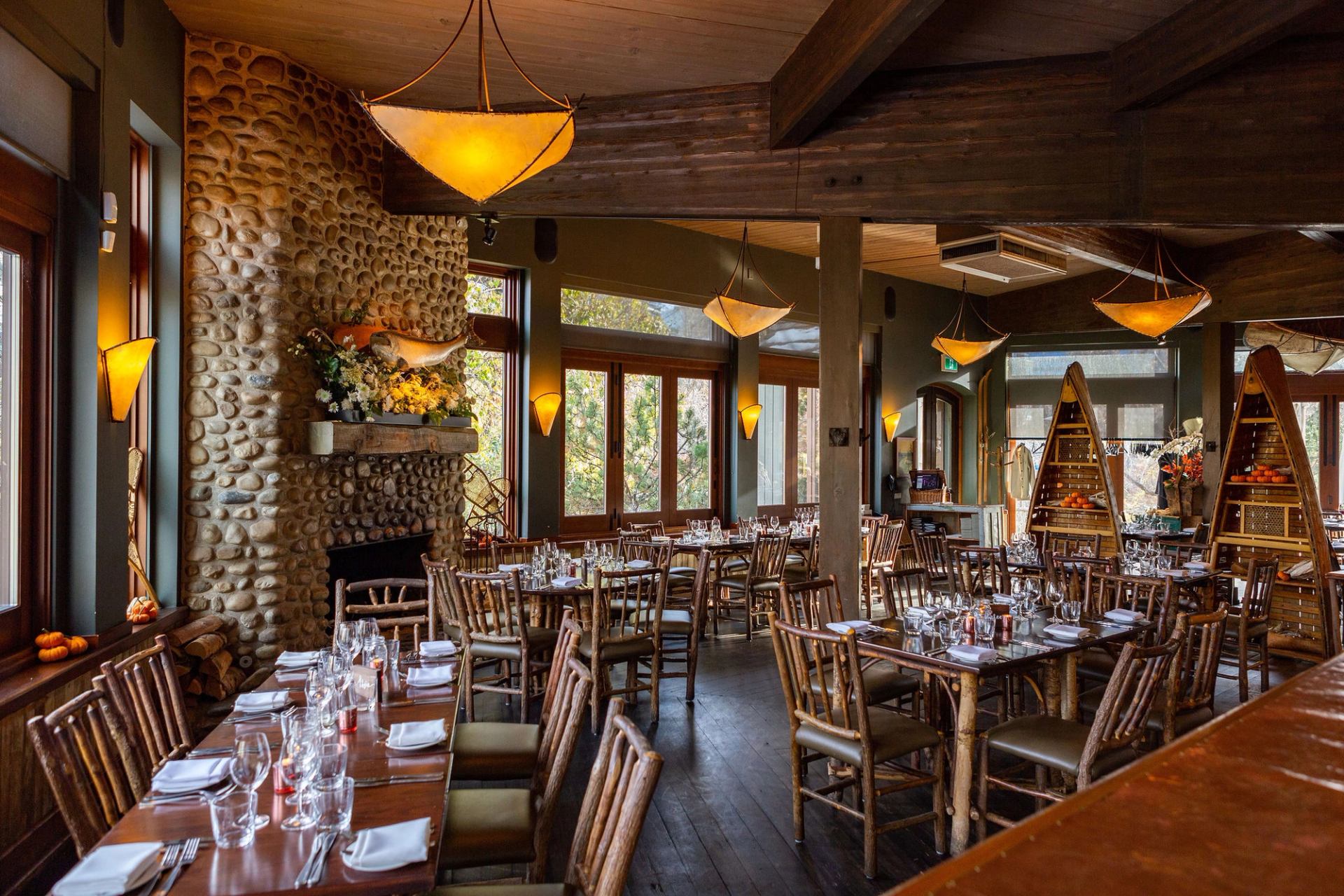 Rustic dining room with set tables, wooden chairs, stone fireplace and pendant lights.
