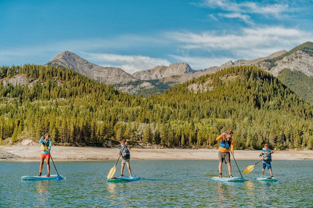 Four people paddleboarding on a lake with forest and mountain backdrop under clear skies.