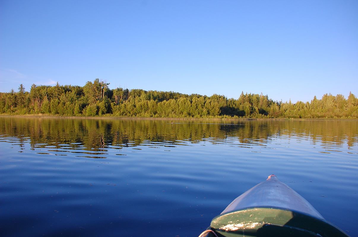 View of one of the Chain Lakes from a kayak, with the bow visible in the foreground and calm water ahead.