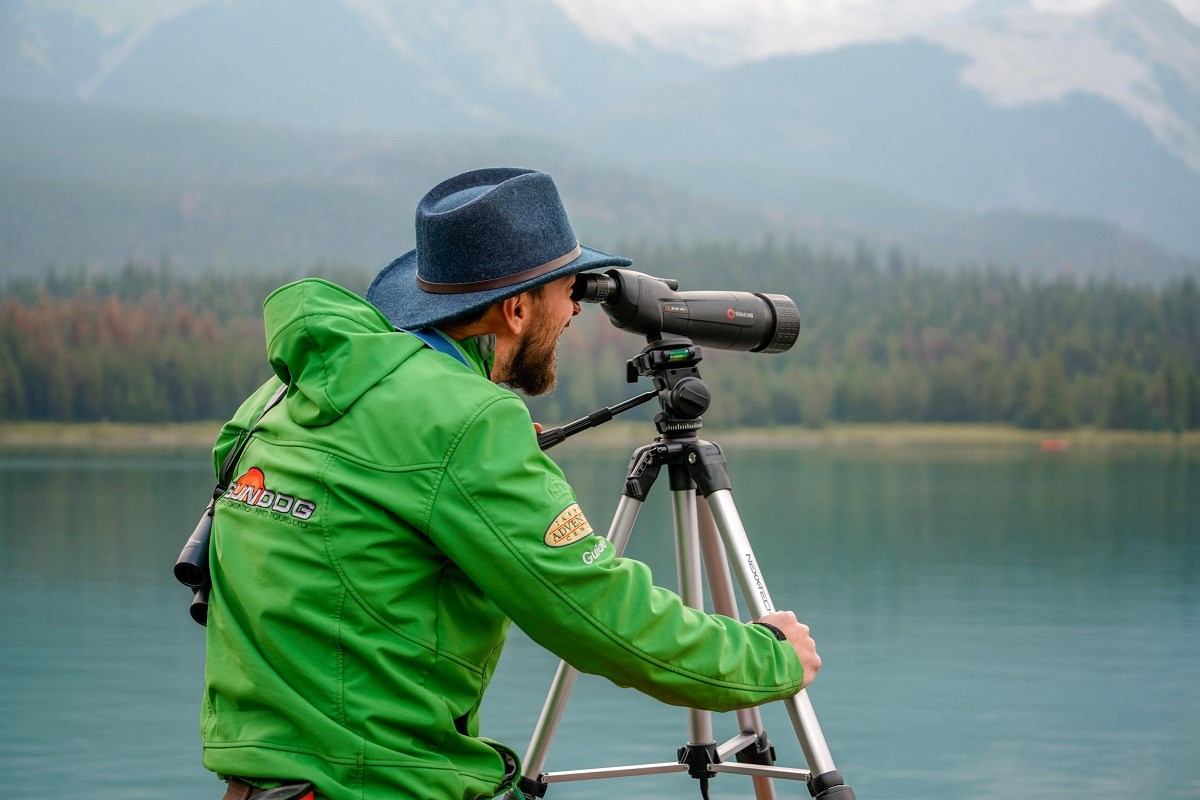 Person in green jacket using spotting scope on tripod beside calm lake and forested mountains