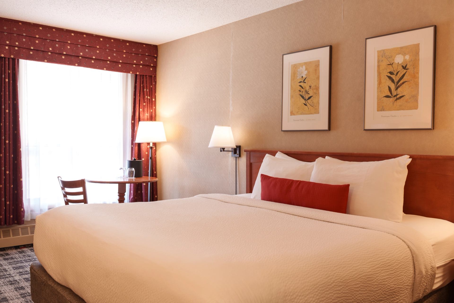 Hotel room with king bed, red cushion, table by window, and framed floral art.