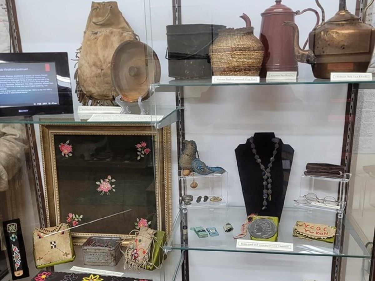 A museum display case with baskets, jewelry, embroidered items, and historical artifacts.
