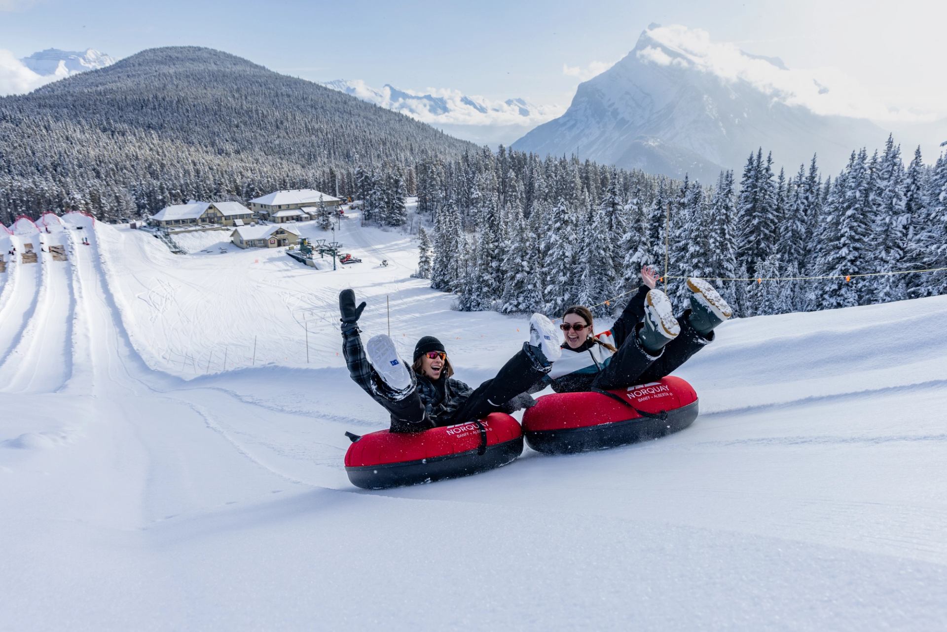 Two people joyfully snow tubing down a hill at a snowy mountain resort.