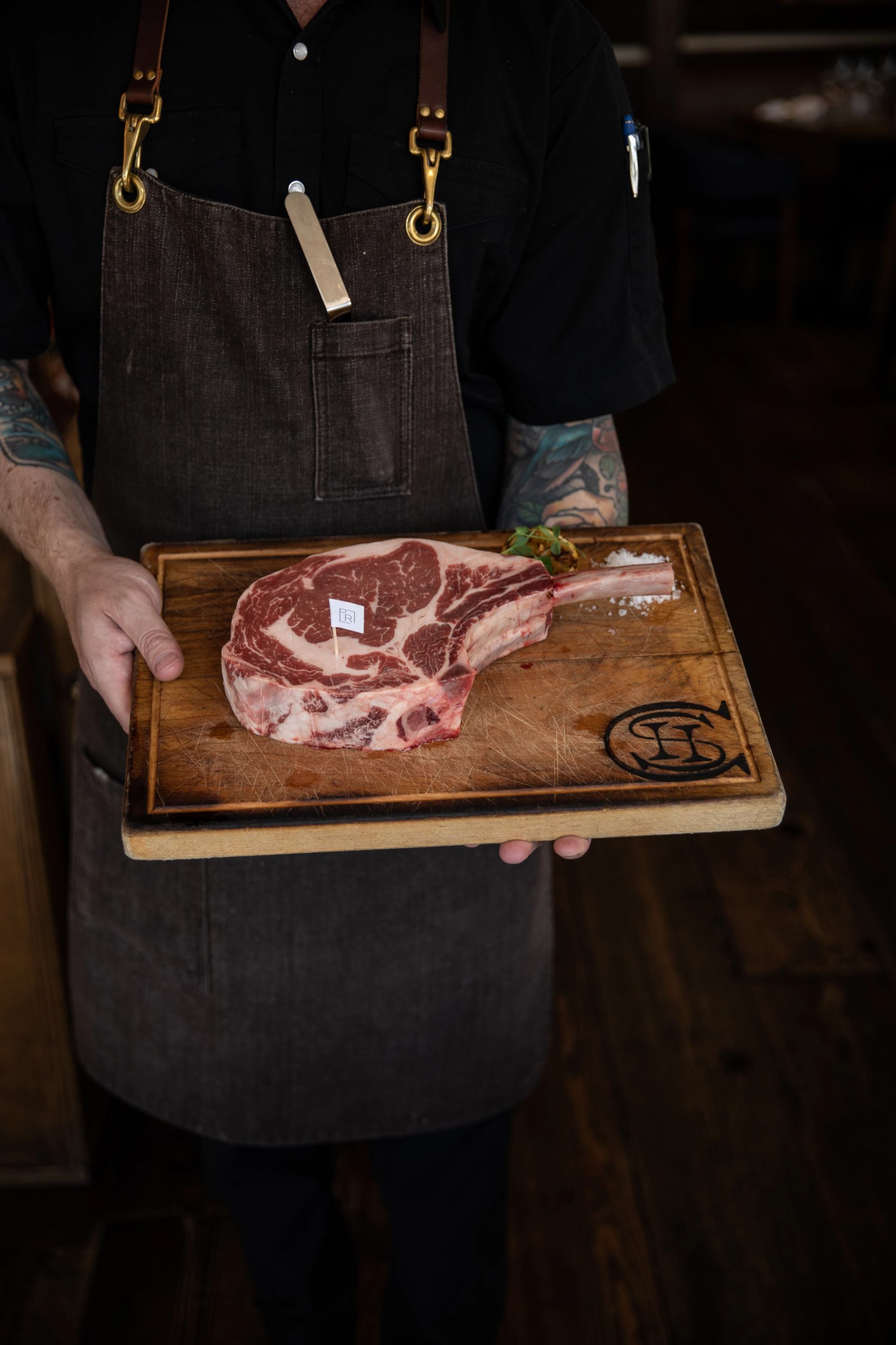 A chef in a dark apron holds a wooden board with a raw tomahawk steak, coarse salt, and a small garnish.