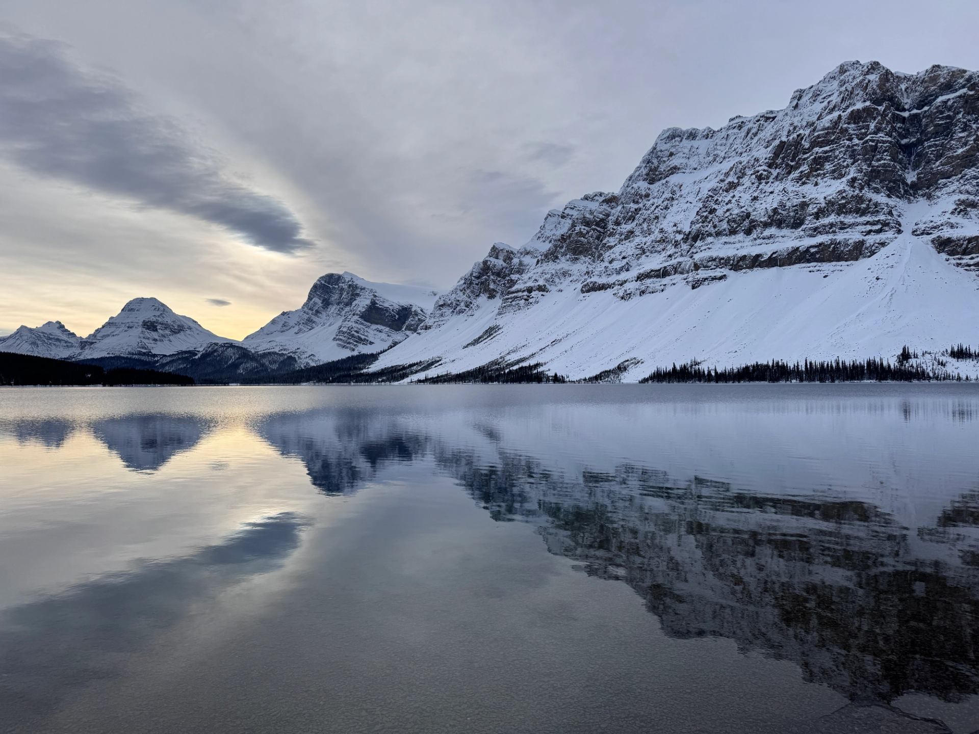 Snow-covered mountains reflected in a calm alpine lake under a cloudy sky.