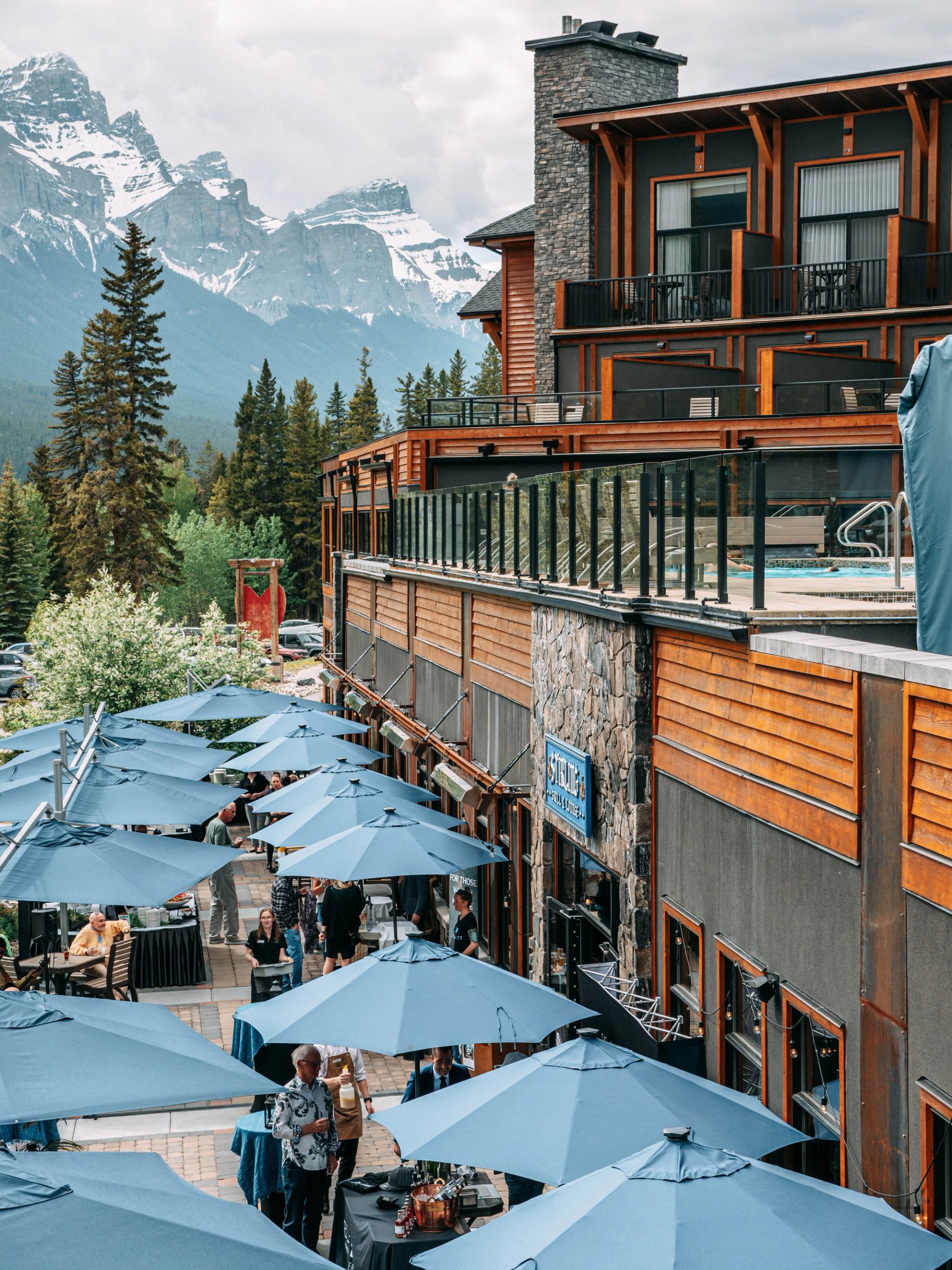 Mountain lodge patio with blue umbrellas, people dining, and snow-covered peaks behind.