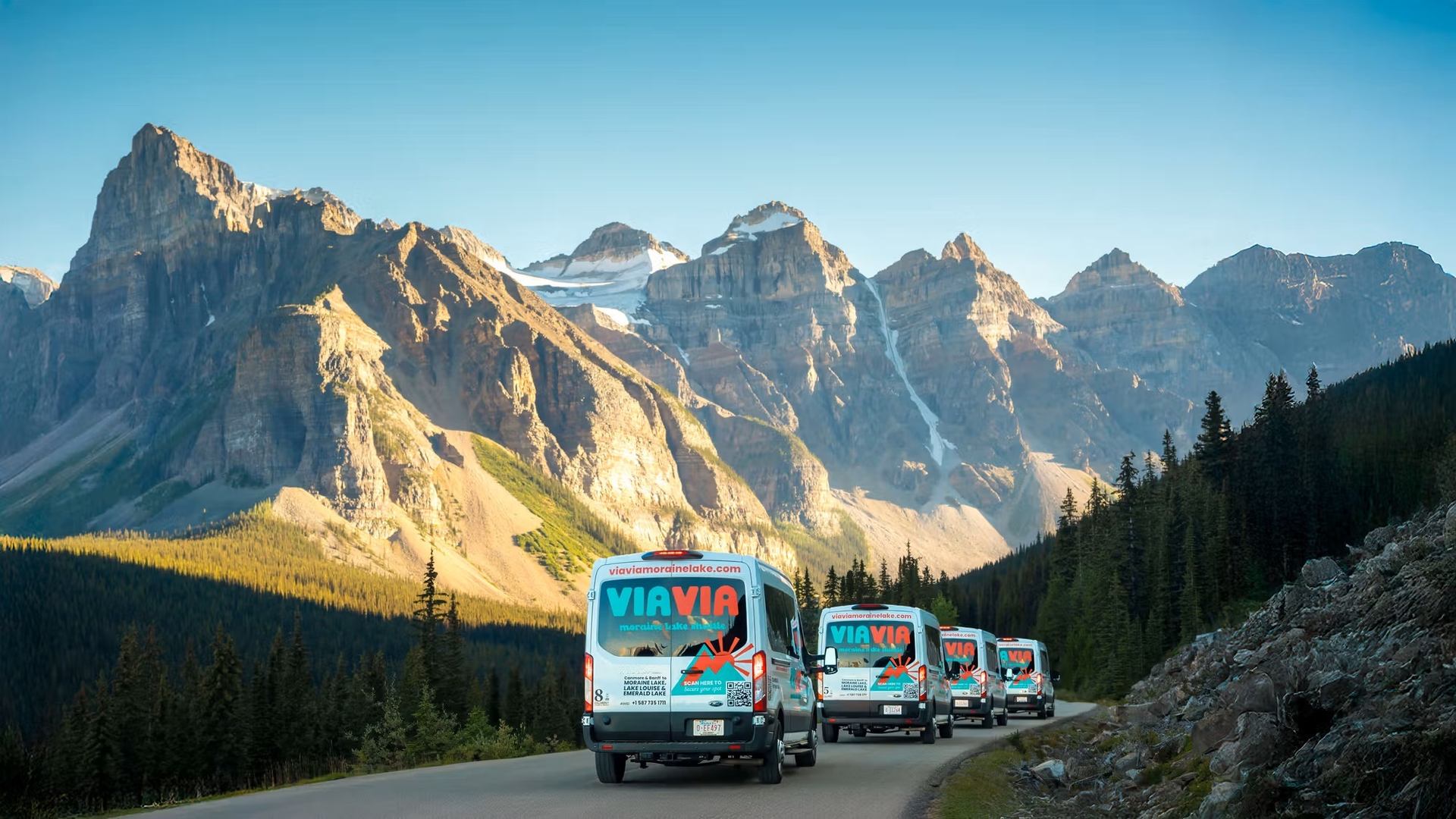 Vans on mountain road with forest, rocky terrain, and snow-capped peaks in background.
