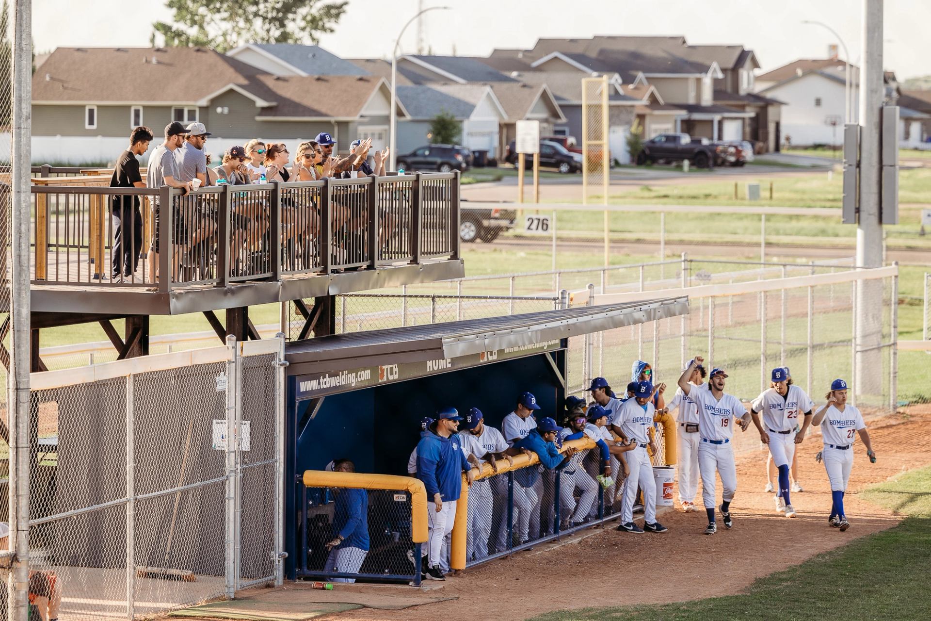 Baseball team gathered in the dugout with fans watching from the viewing deck above.