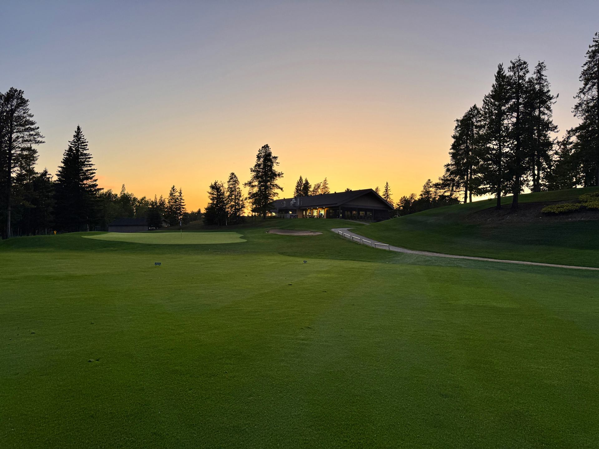 Sunset view of The Dunes Golf & Winter Club with rolling greens, clubhouse, trees, and a glowing sky.