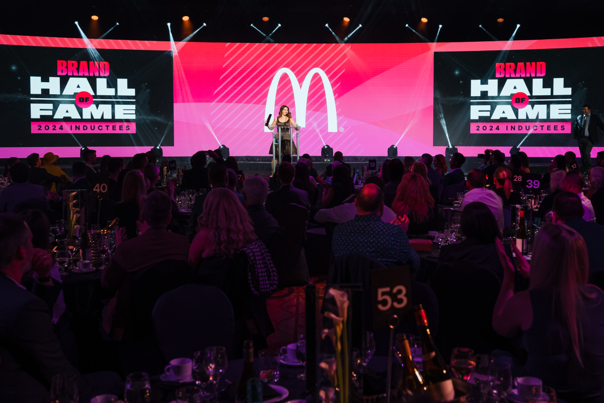 Conference stage with speaker at podium, “Brand Hall of Fame 2024 Inductees” backdrop and audience i.