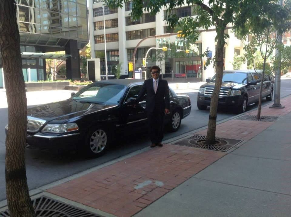 Man in suit next to black limo on city sidewalk, another vehicle and buildings in background.