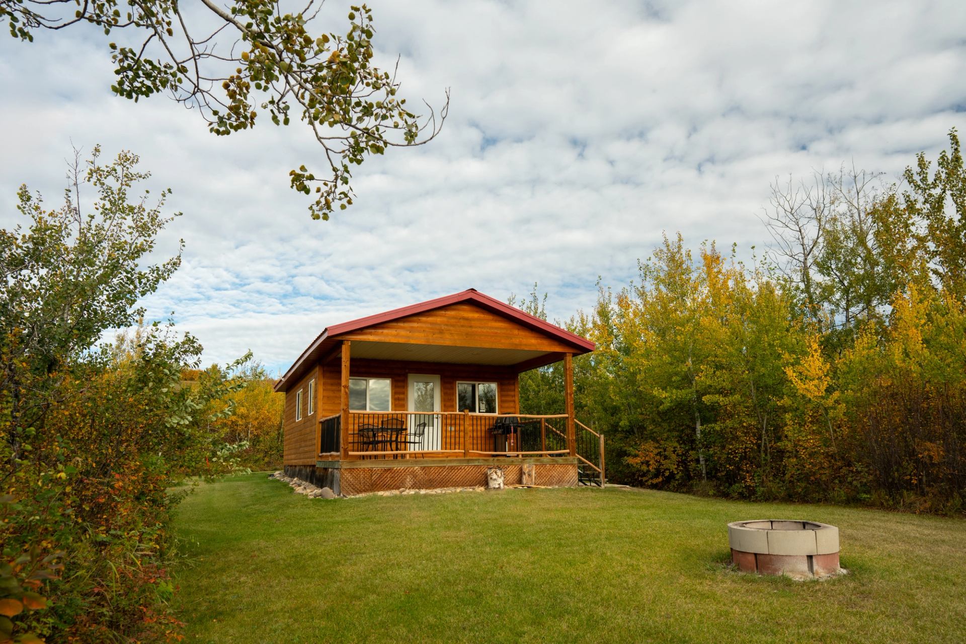 Wooden cabin with red roof, trees, grassy lawn, and fire pit.