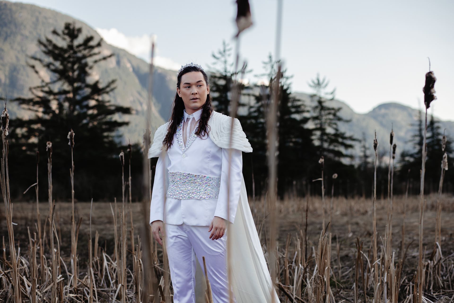 Performer in white outfit standing outdoors with mountains and pine trees in background.