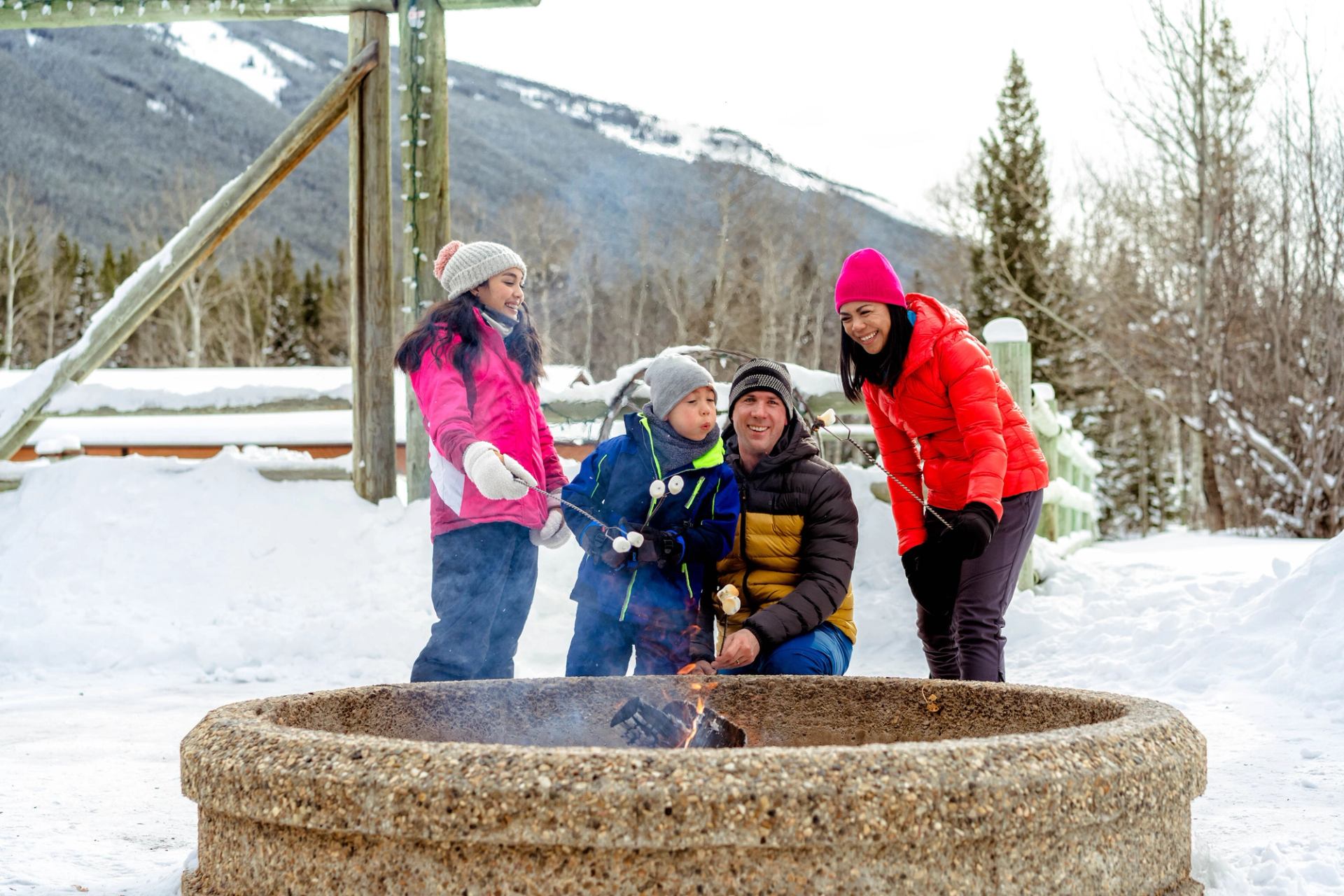 Family gathered around an outdoor fire pit roasting food in a snowy Rocky Mountain setting.