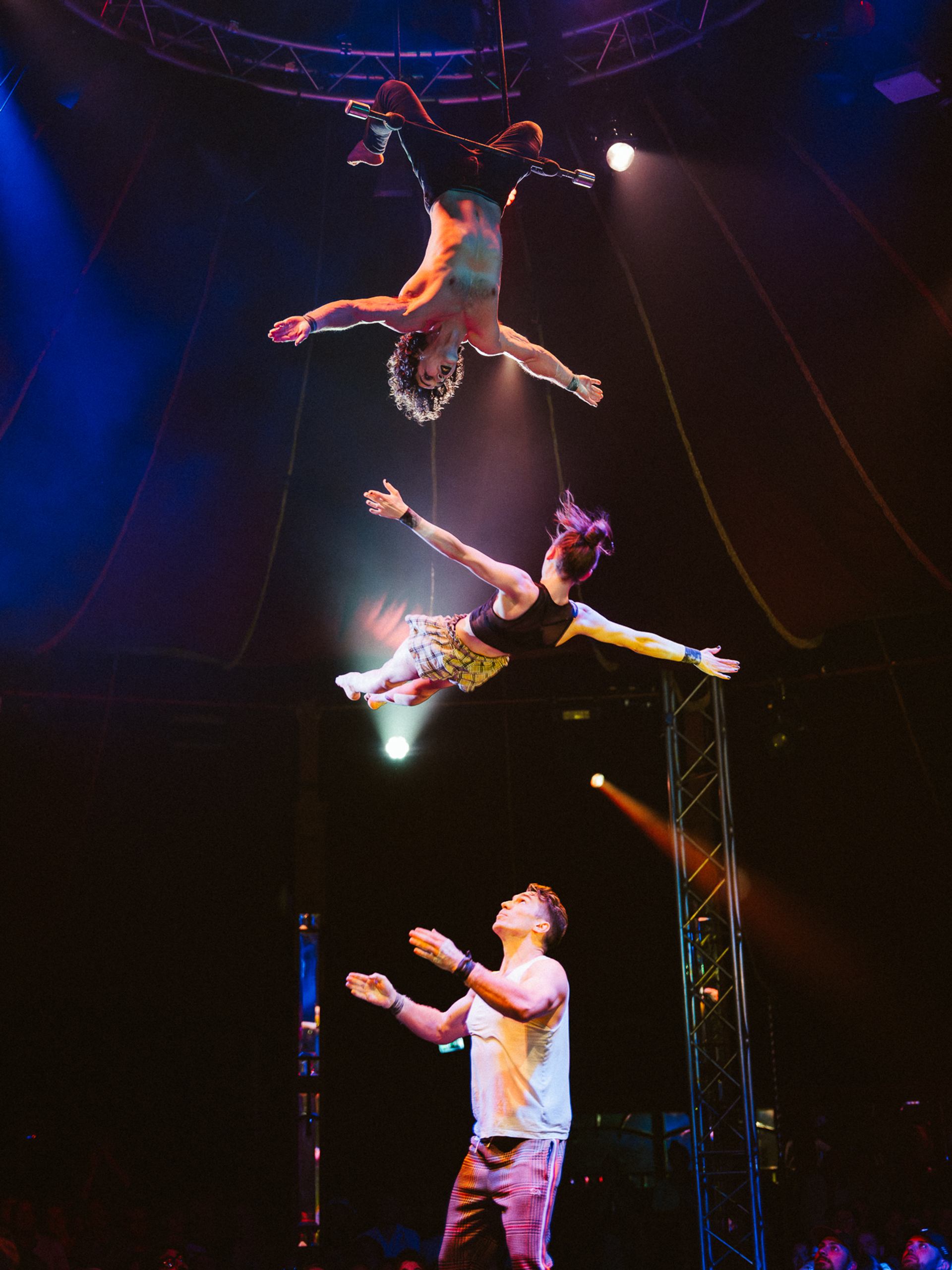 Circus acrobats perform aerial stunt above stage, with spotlights and audience below.