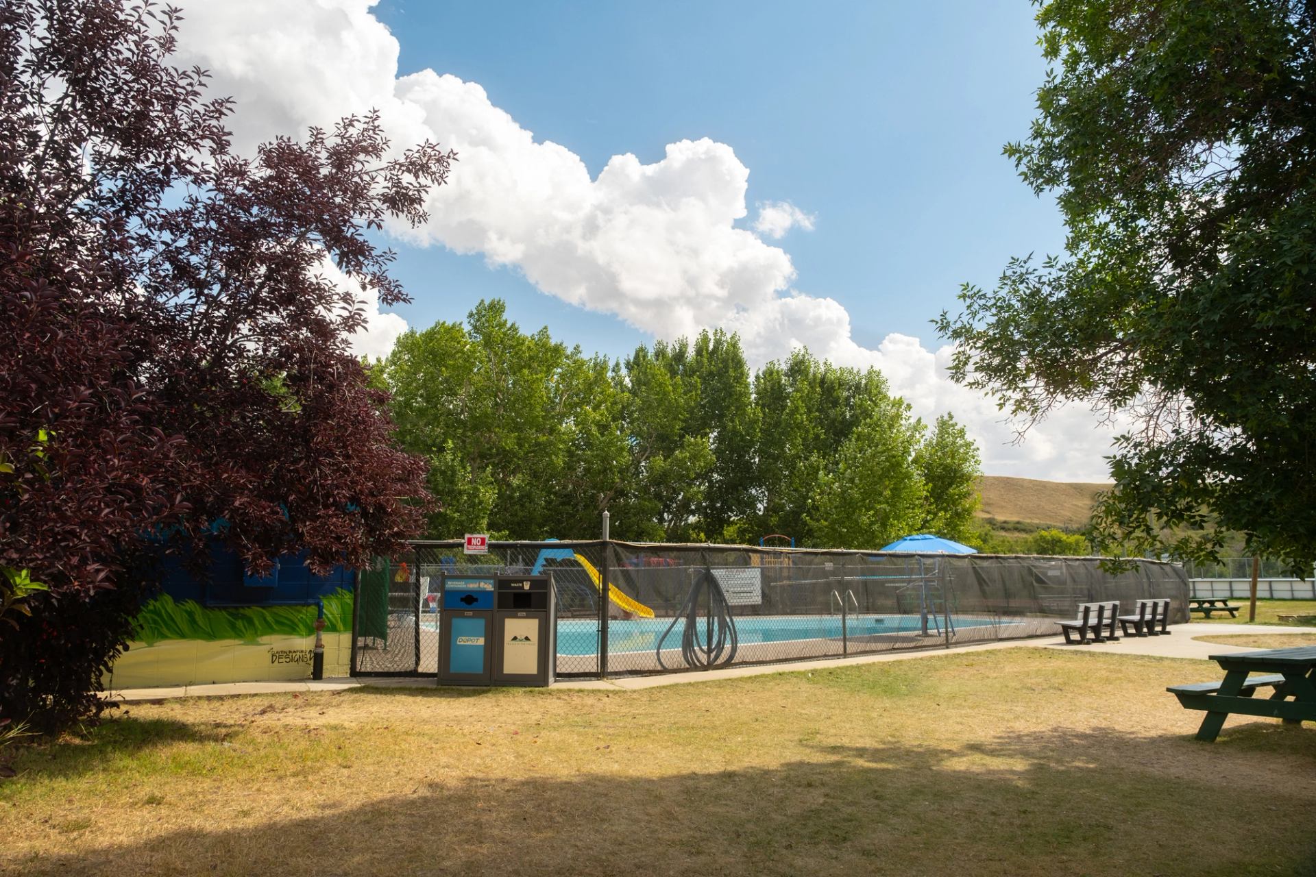 Swimming pool with a slide, fenced in, surrounded by trees and picnic tables under a blue sky.