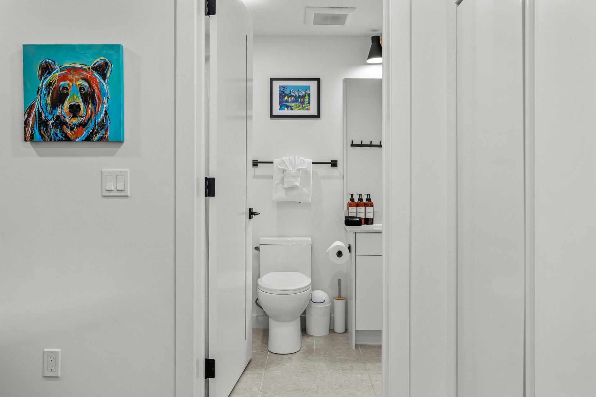 White bathroom with toilet, vanity, wall art and colourful bear painting in hallway.