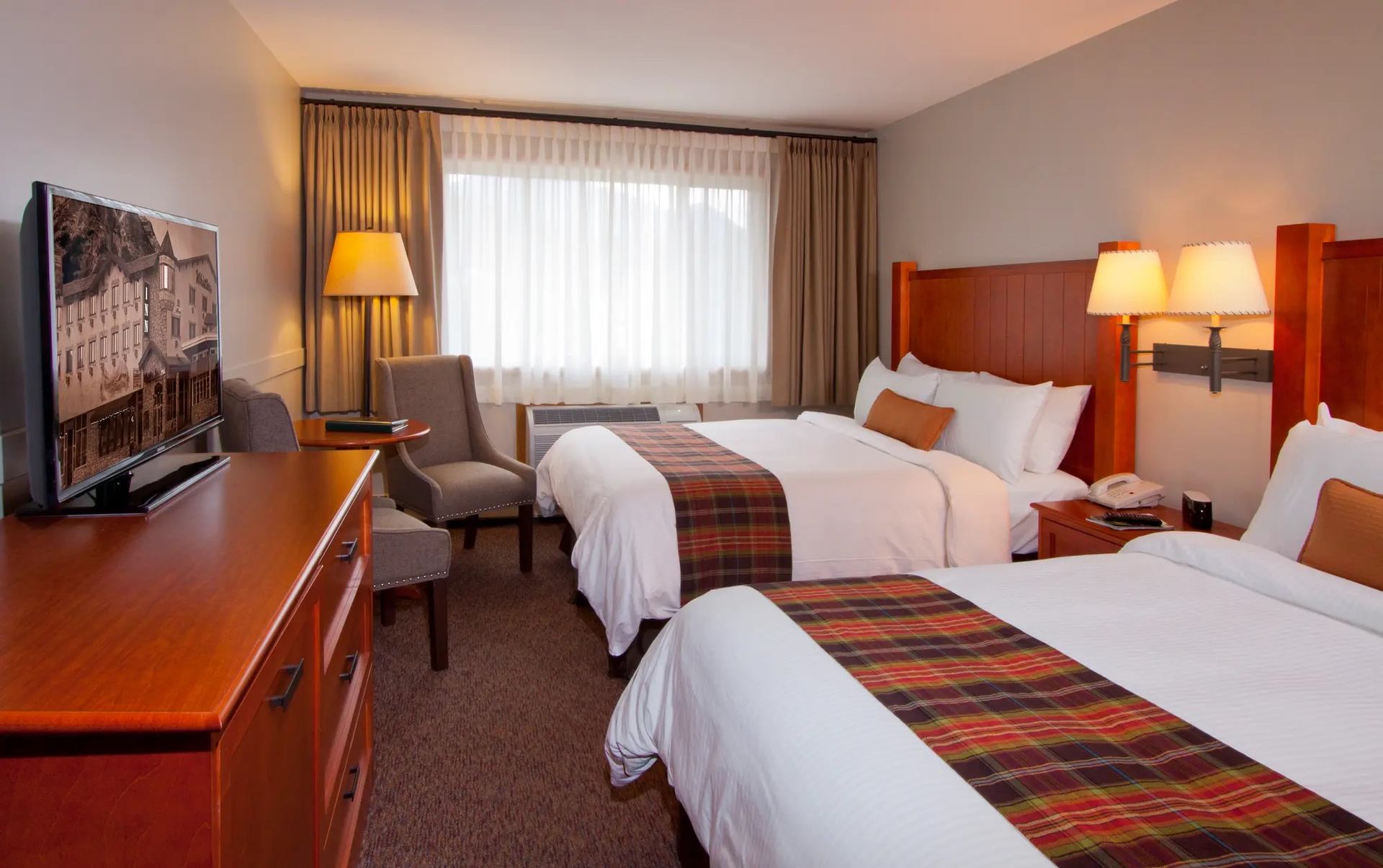 Cozy hotel room with two beds, plaid blankets, warm lighting, and window seating.