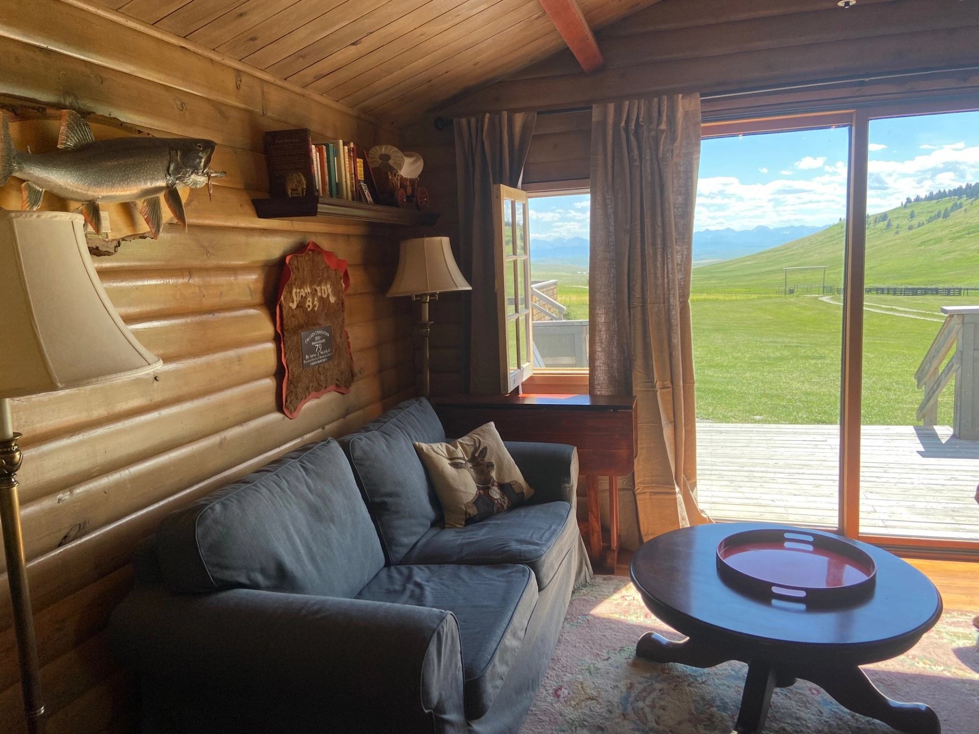 Rustic living room with log walls, gray sofa, wall-mounted fish décor, and glass doors to green hills.