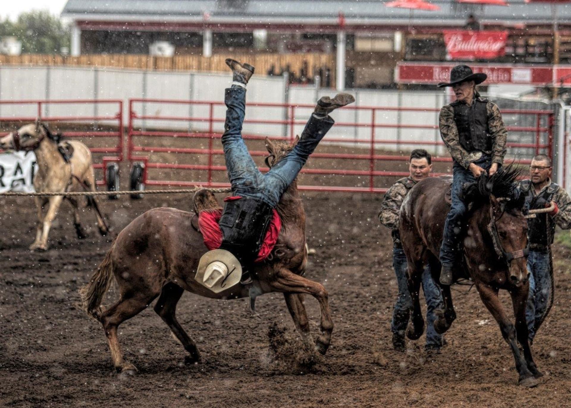 Rodeo rider flips off bucking horse in muddy arena as others approach on horseback.
