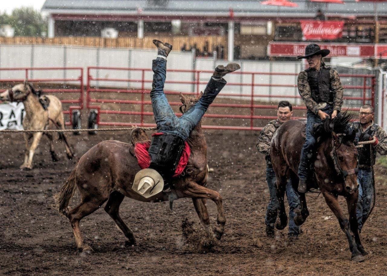Rodeo rider flips off bucking horse in muddy arena as others approach on horseback.