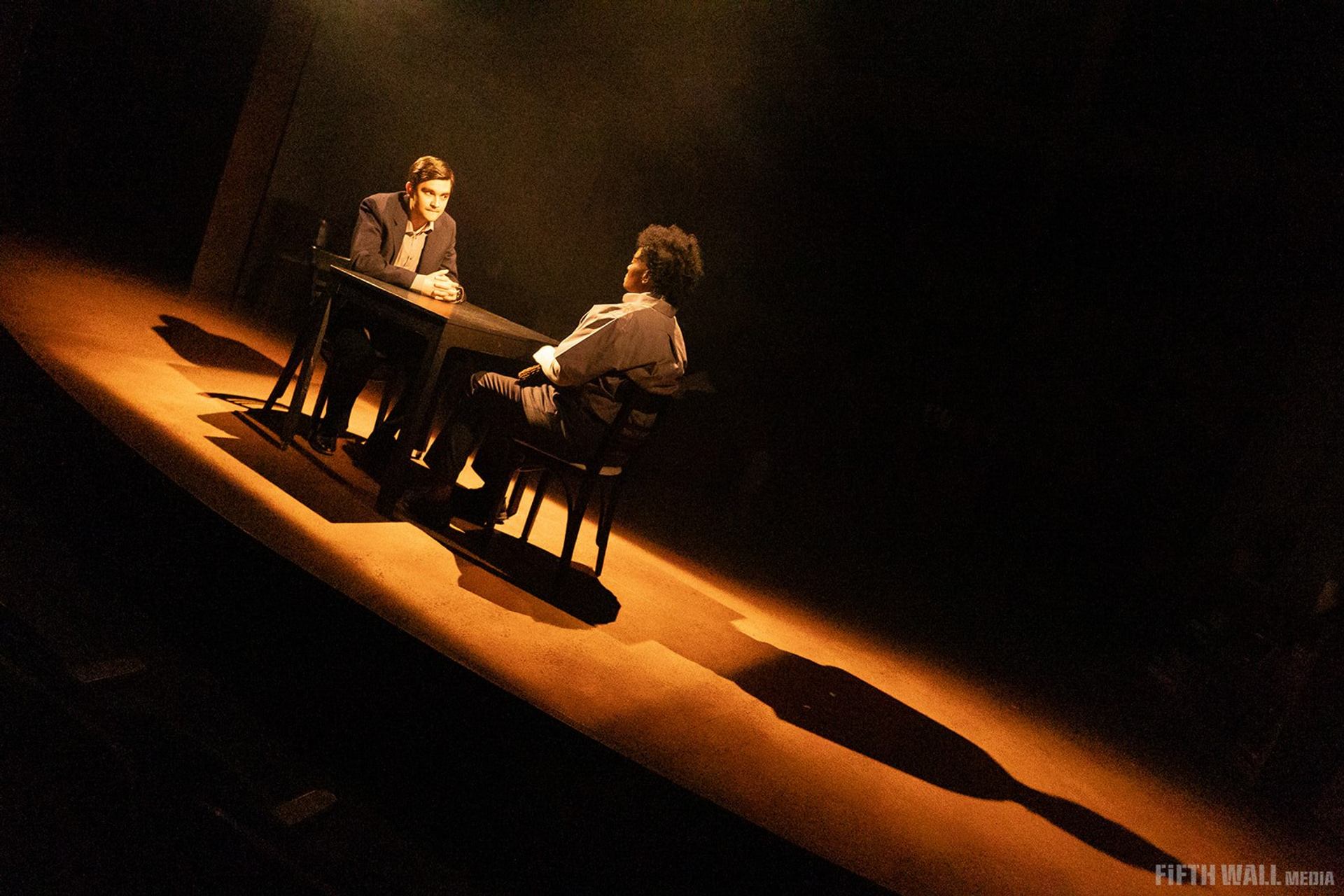 Stage scene with two people seated at a table under dramatic spotlight.