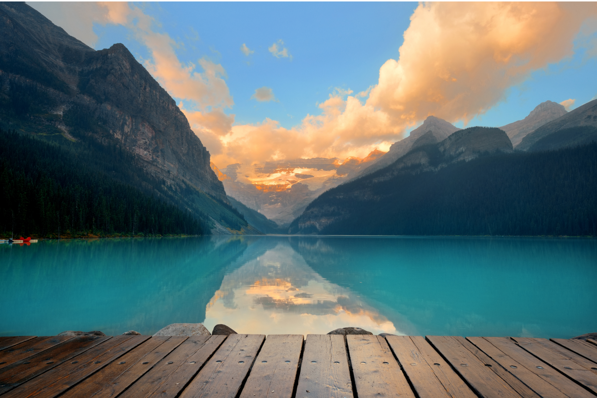 Wooden dock overlooking turquoise water and mountains under a colorful sunrise sky.