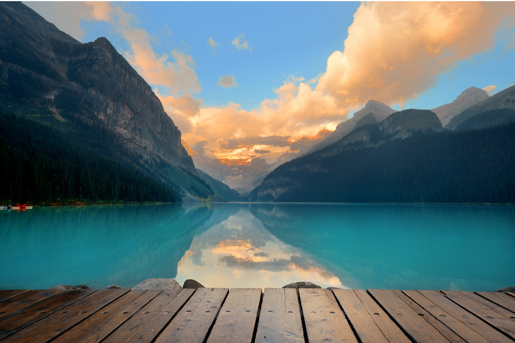 Wooden dock overlooking turquoise water and mountains under a colorful sunrise sky.