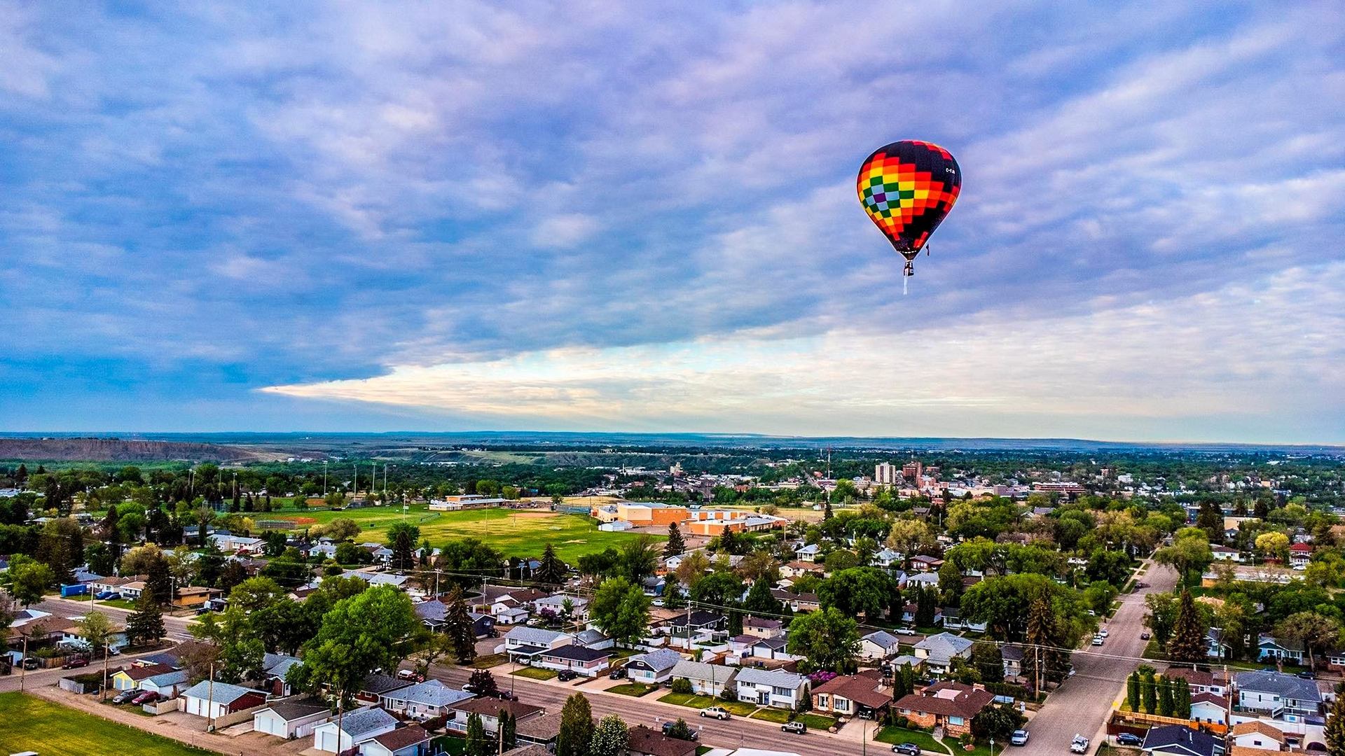 A colorful hot air balloon floating above a city with trees and houses under a cloudy sky.