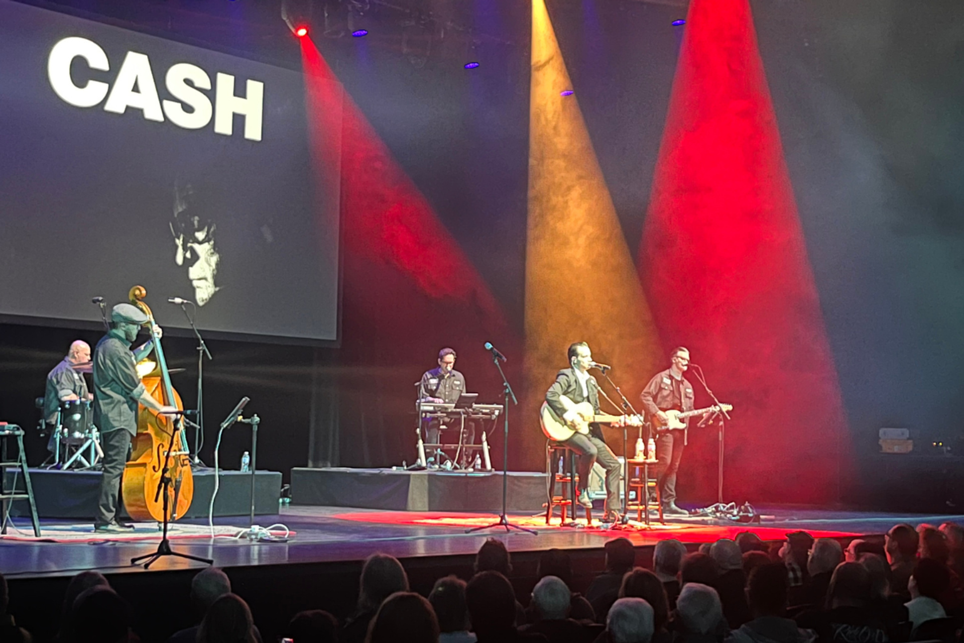 Band performs onstage under coloured spotlights with large CASH sign and portrait backdrop.