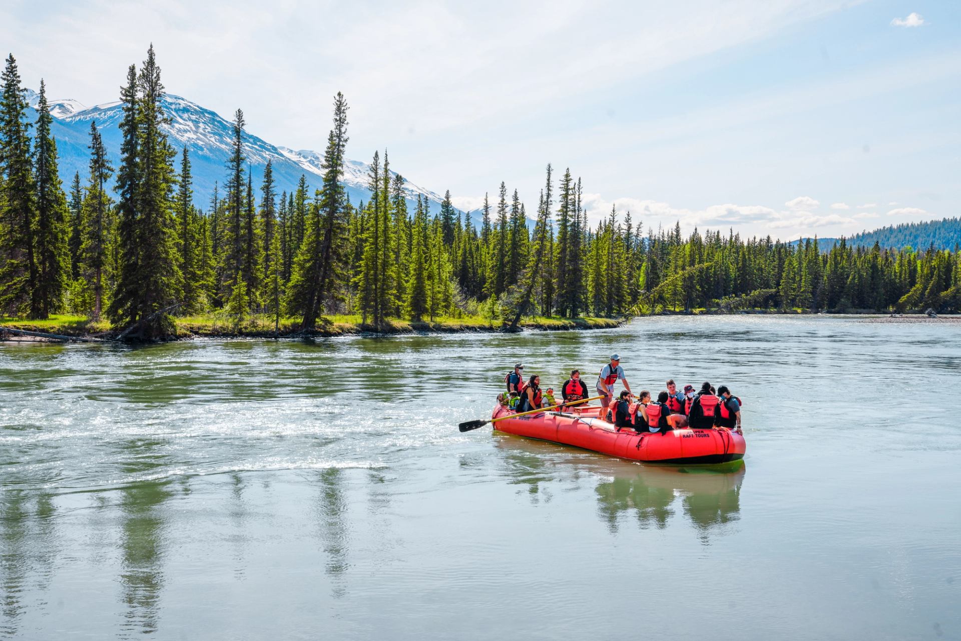 A red raft carries guests along a wide river with pine trees and mountains.