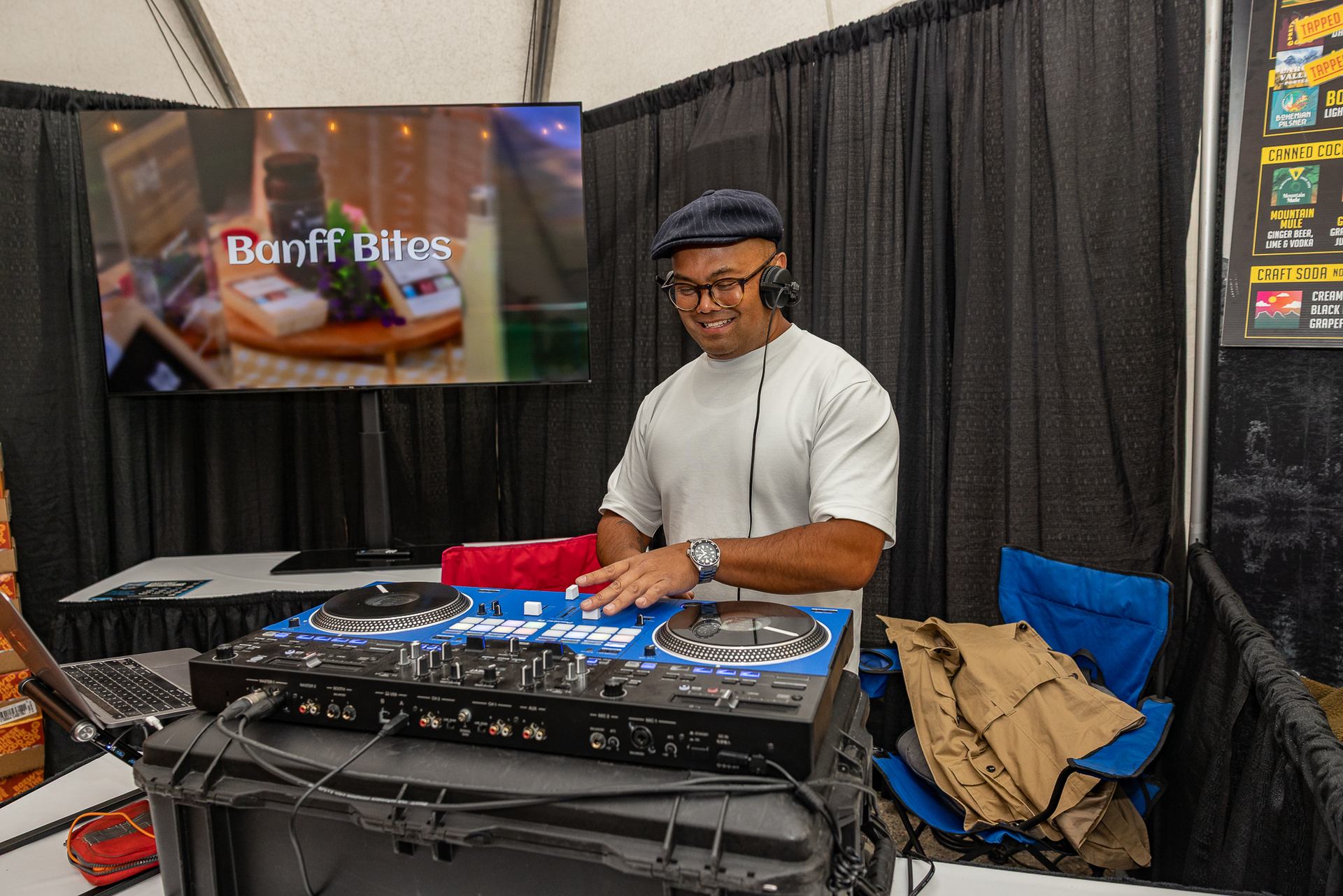 DJ wearing headphones mixes on turntables beside Banff Bites display screen at booth.