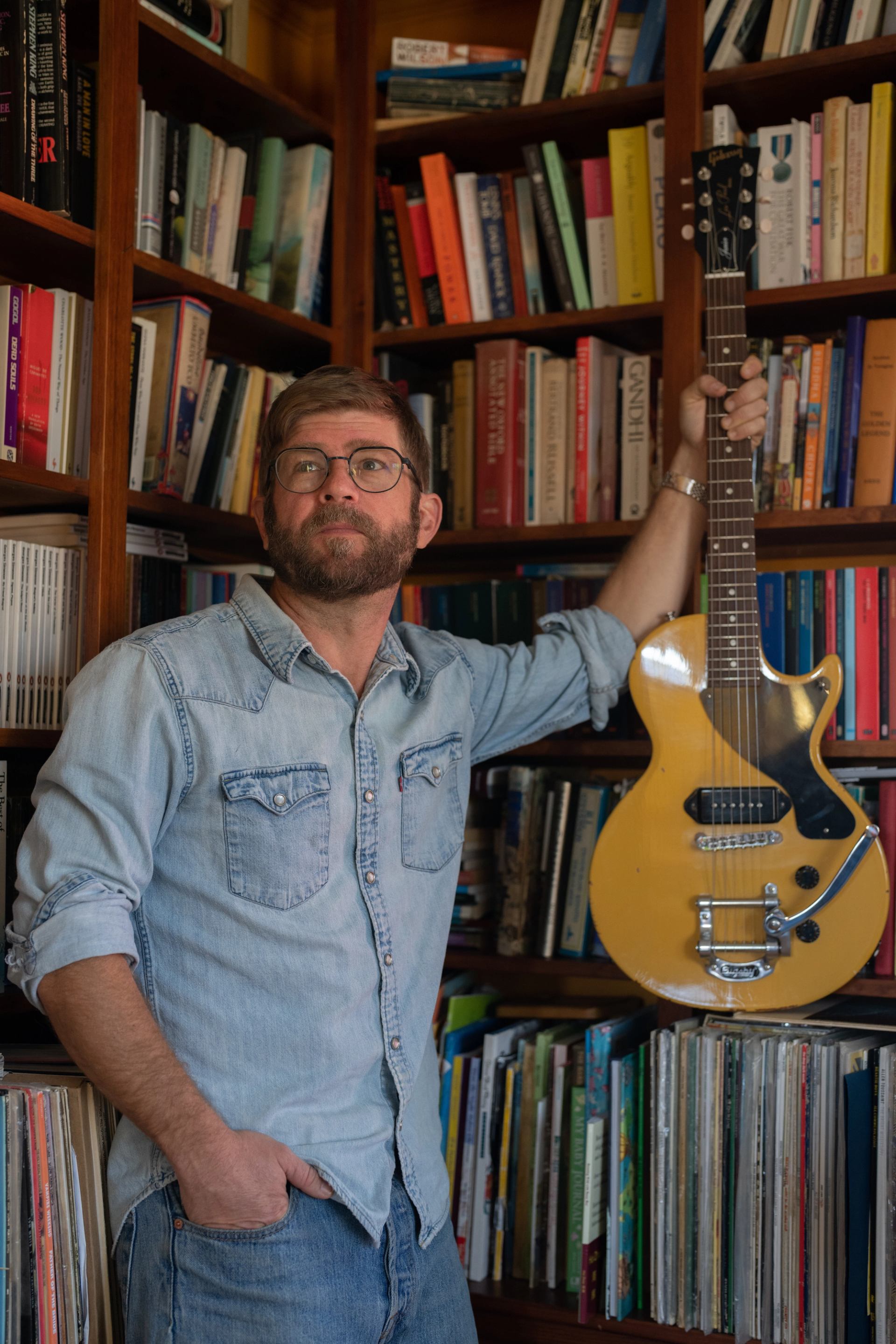 A bearded man in a denim shirt and glasses holds a yellow electric guitar, standing in front of a large bookshelf.
