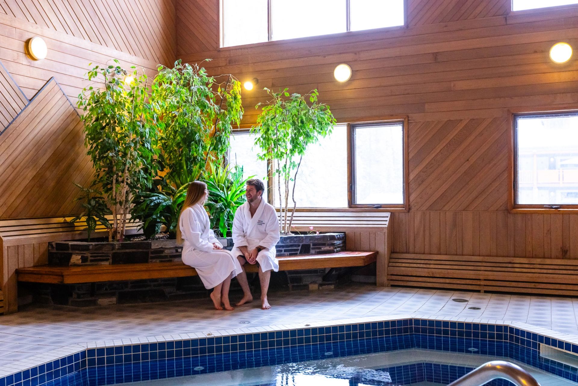 A couple in white robes relaxing on a wooden bench in a bright, plant-filled indoor hot tub area.