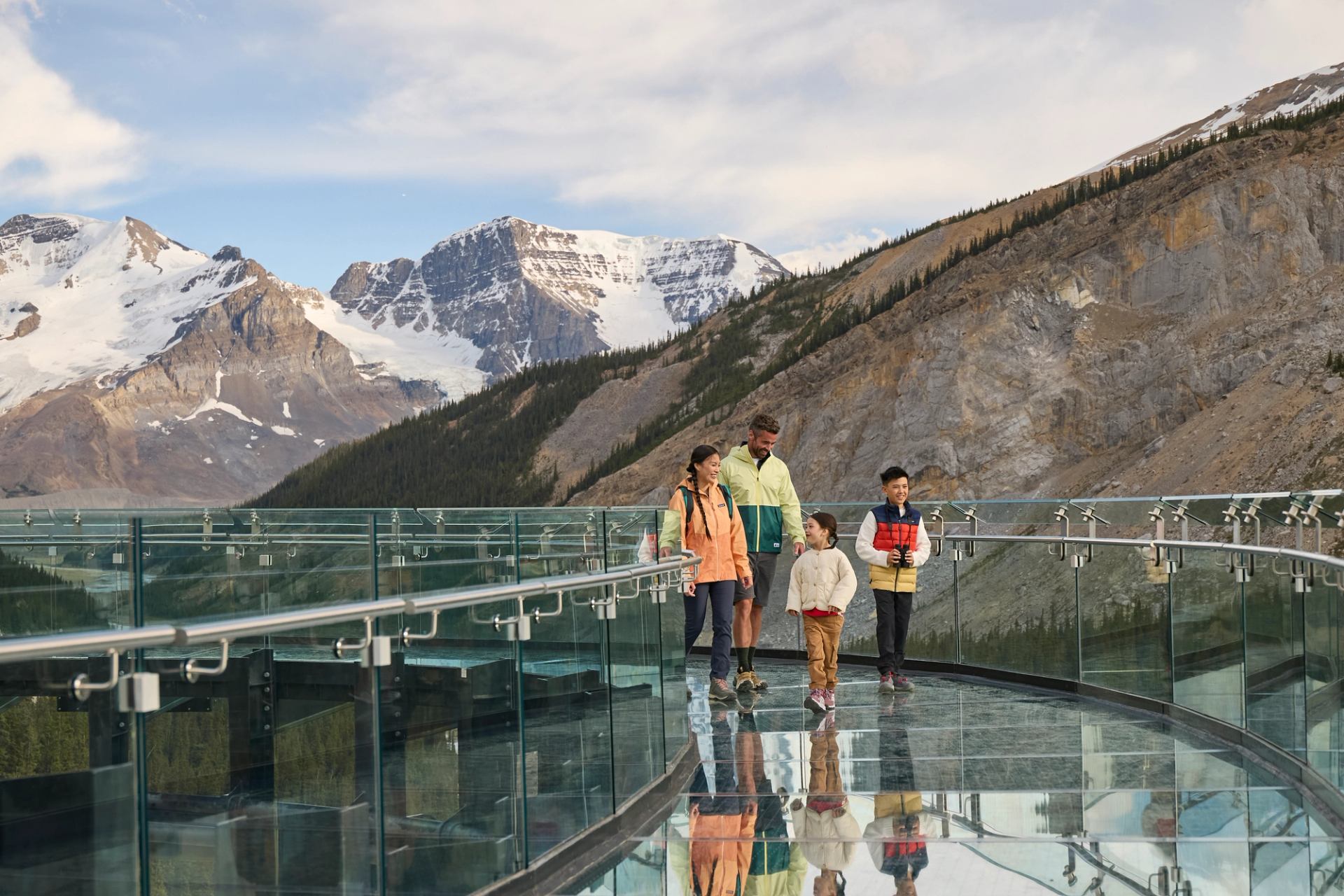 People walking on a glass observation walkway with snowy mountains and rocky cliffs behind.