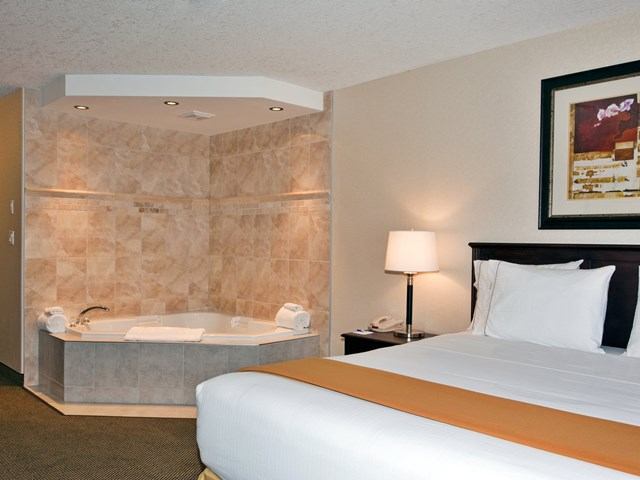 A hotel suite showcasing a neatly made bed with white linens and a tan throw runner next to a private corner jetted bathtub with beige tiled walls.