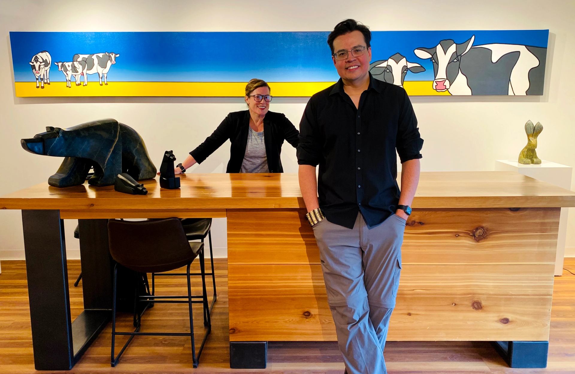 Art gallery interior with two people at wooden counter and colourful cow painting.