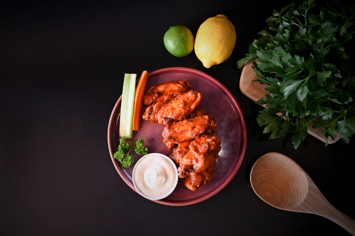 Buffalo wings with dipping sauce, celery, carrots, and fresh garnish on a dark plate.