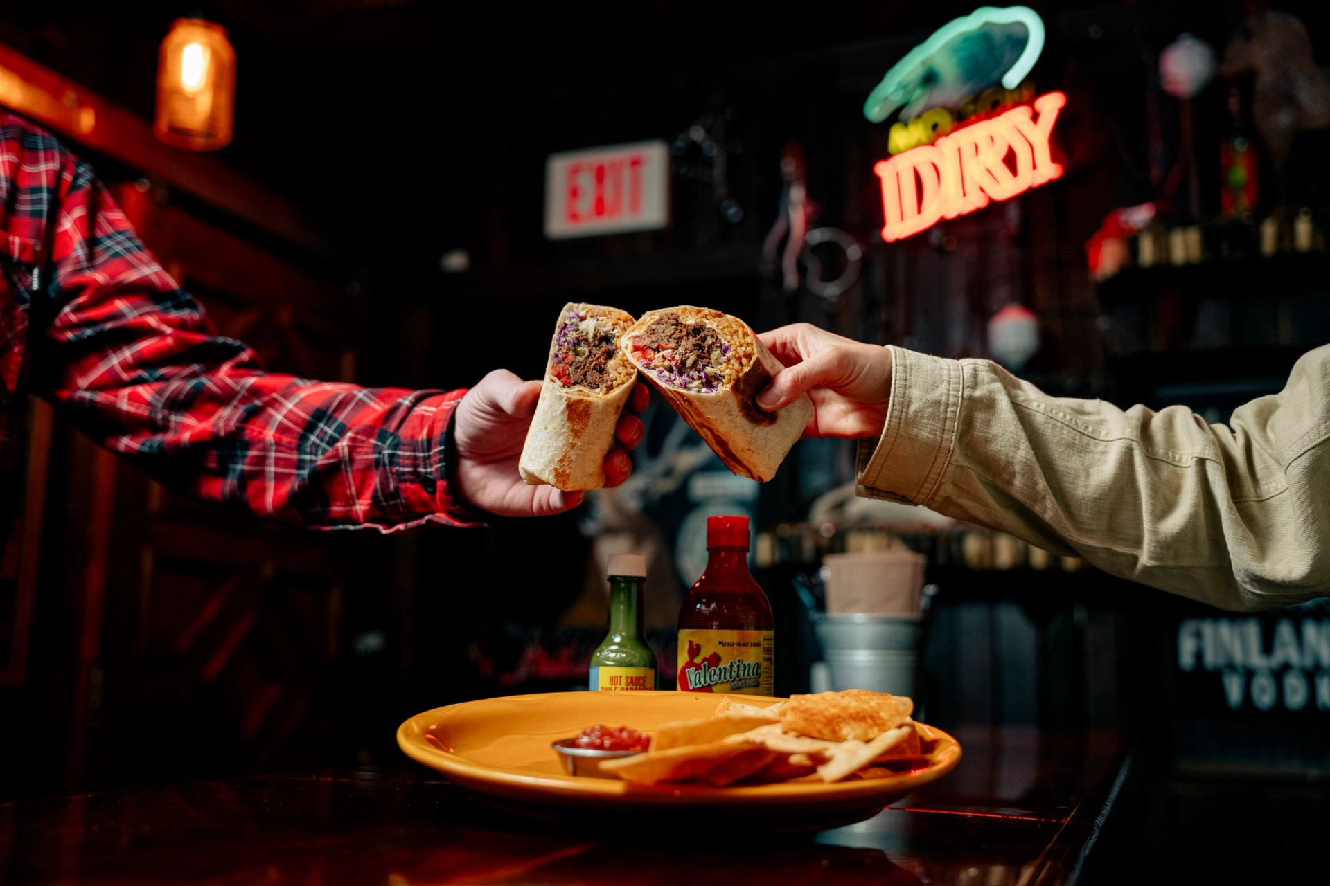 Two hands clinking halves of burritos together over a plate of chips and salsa at a bar.