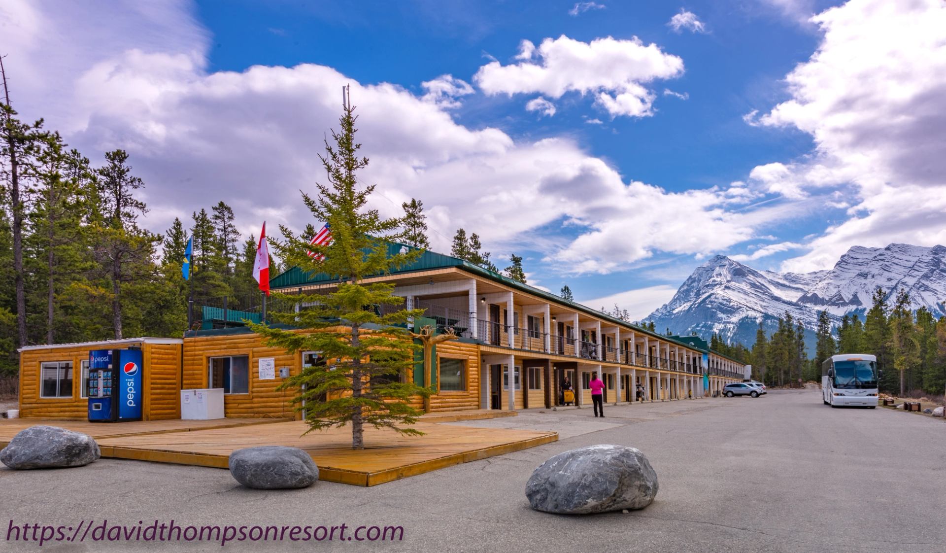 Log resort building with parked bus and snow-capped mountains under cloudy blue sky.