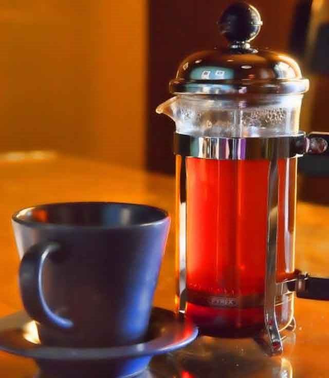 French press with red tea or coffee and a cup on a wooden table in warm lighting.