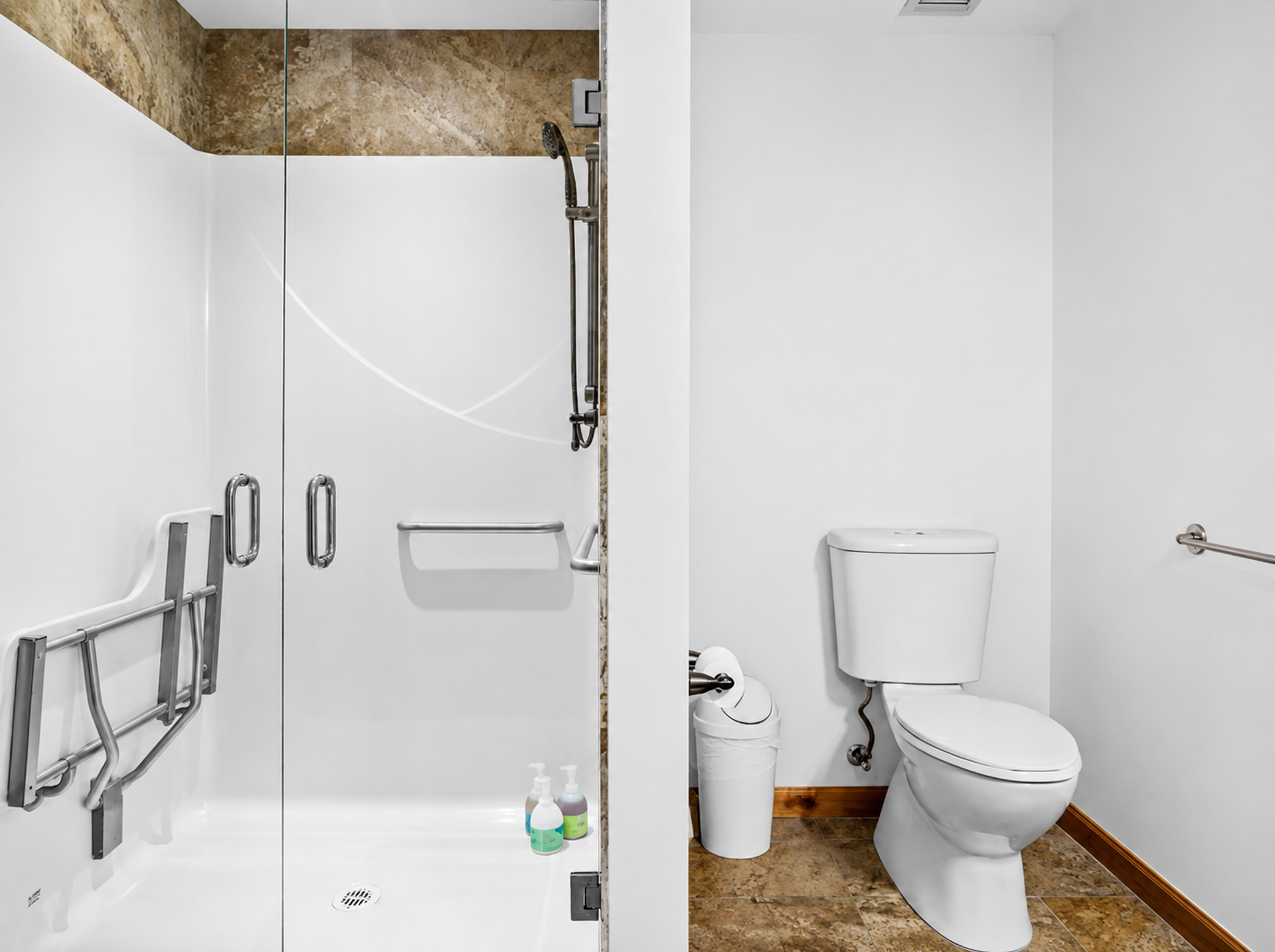 Accessible bathroom with glass shower, grab bars, toilet and waste bin on tile floor.