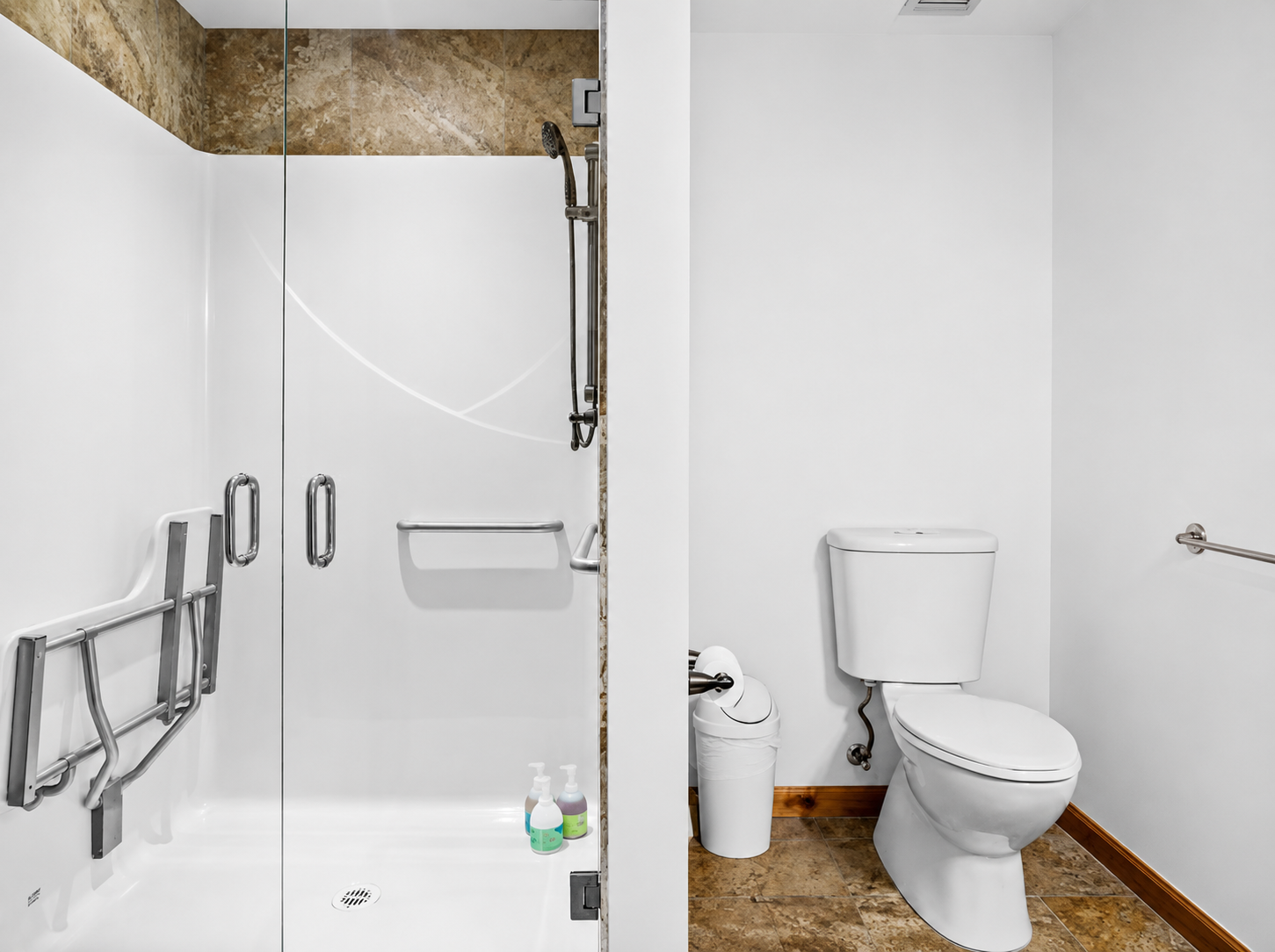 Accessible bathroom with glass shower, grab bars, toilet and waste bin on tile floor.