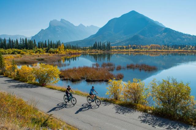 Two cyclists travel along a lakeside road with mountains in the background.