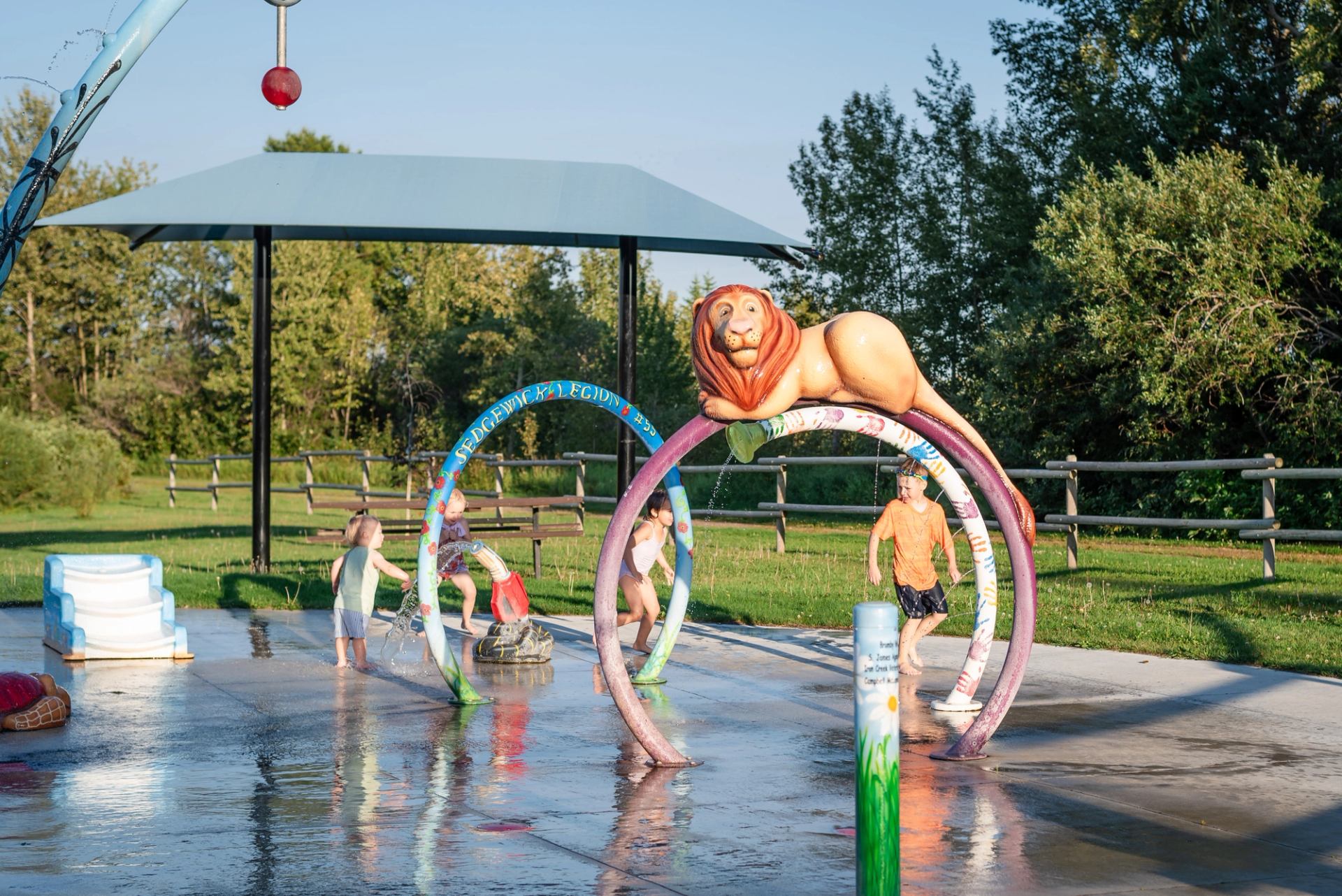 Children run through lion-themed splash park arches with water spraying on wet concrete.
