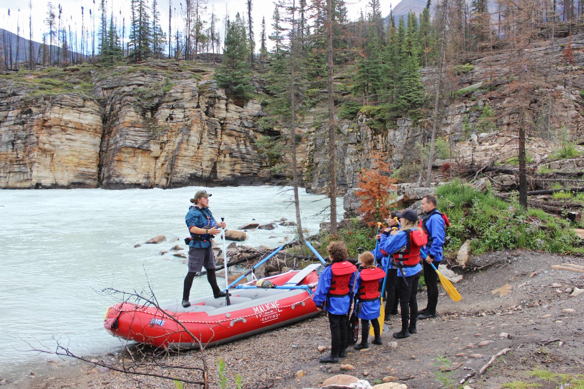 A rafting guide stands on a red raft, addressing a group of people on a rocky riverbank, with cliffs and trees in the background.