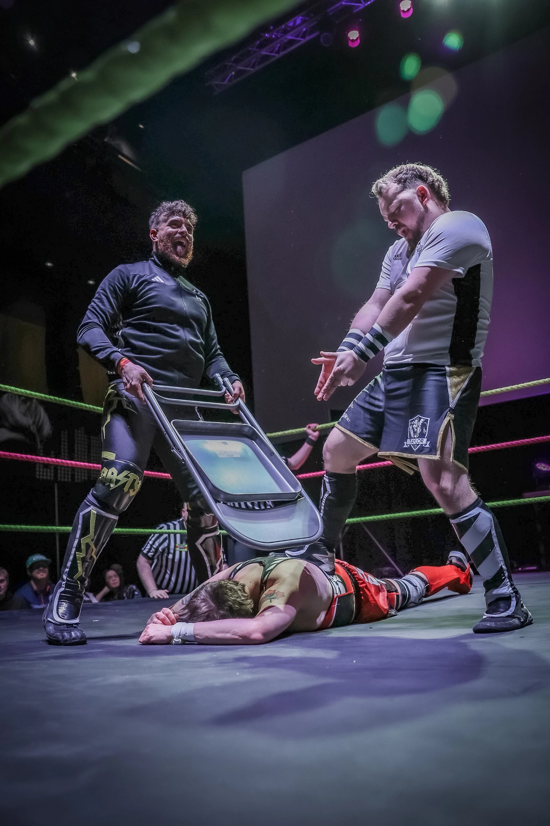 Wrestlers hold a folding chair over an opponent lying face down in the ring.